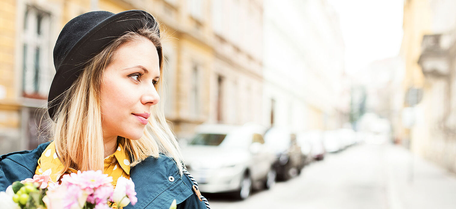 A young woman with blonde hair is wearing a black hat and a blue coat. She is holding a bouquet of flowers and standing on a city street with cars parked along the sidewalk