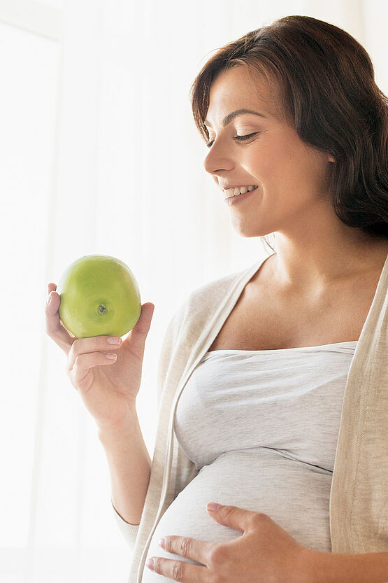 Pregnant woman holding a green apple, smiling and gently touching her belly, standing in a bright room.