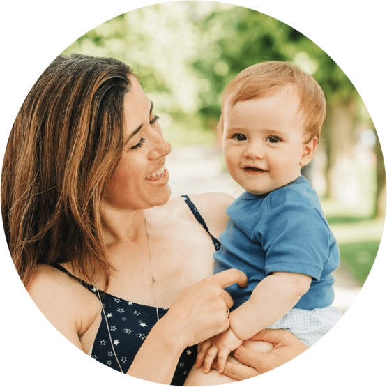 Mother holding her smiling baby, both looking happy, in a sunny outdoor setting with green trees in the background.