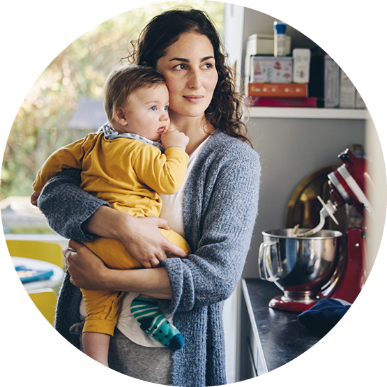 Mother holding her baby, who is sucking on their thumb, in a cozy kitchen setting with a mixer and other kitchen items in the background.