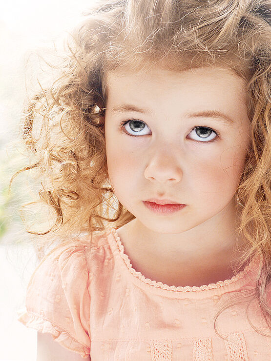 A young girl with curly blonde hair, wearing a light pink dress, looking upward with a slightly distressed expression